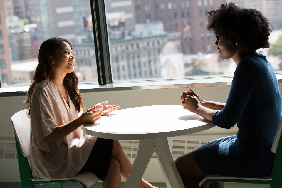 Two people talking across a small table by an office window